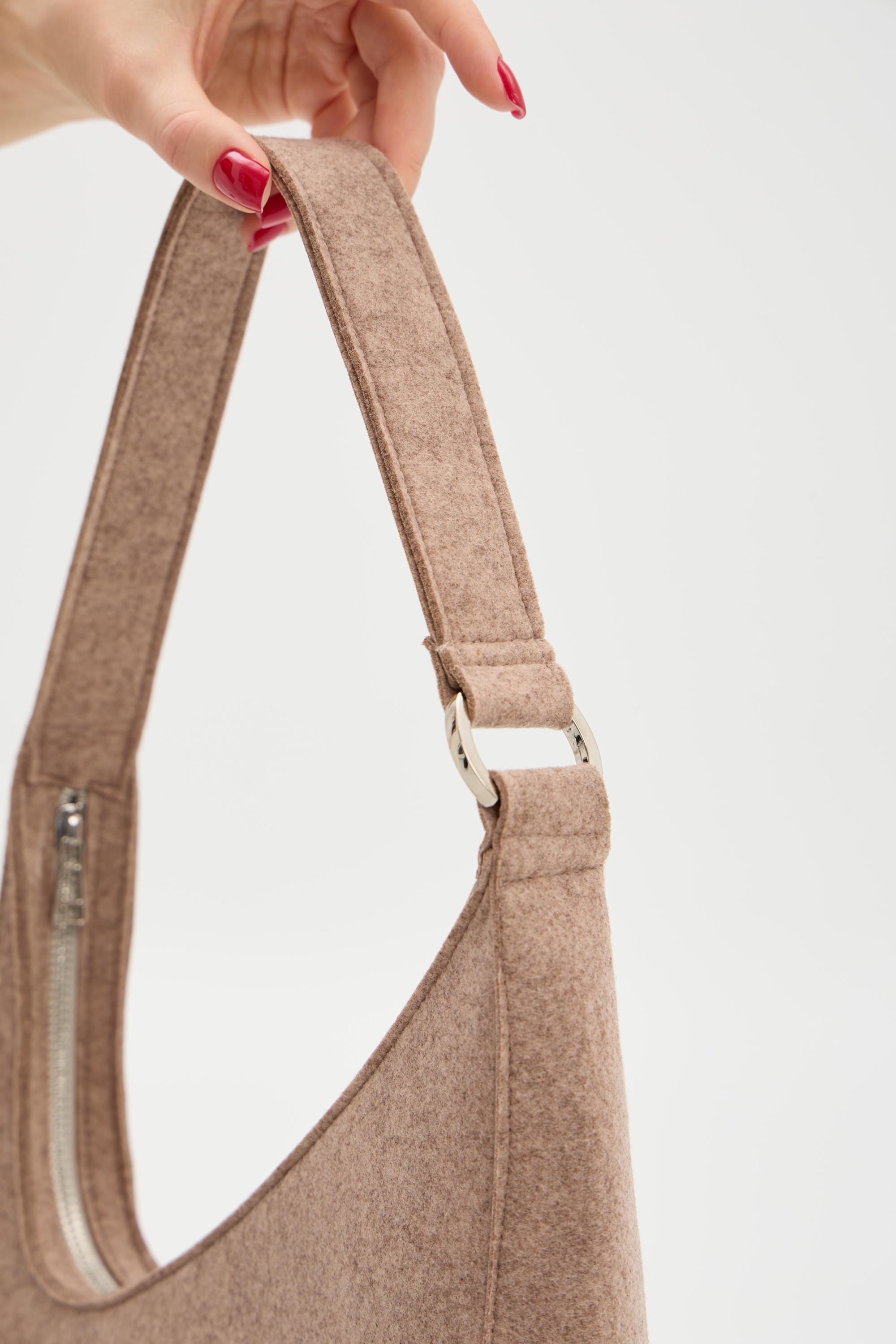Beige shoulder bag held by a hand with red nail polish on a light gray background