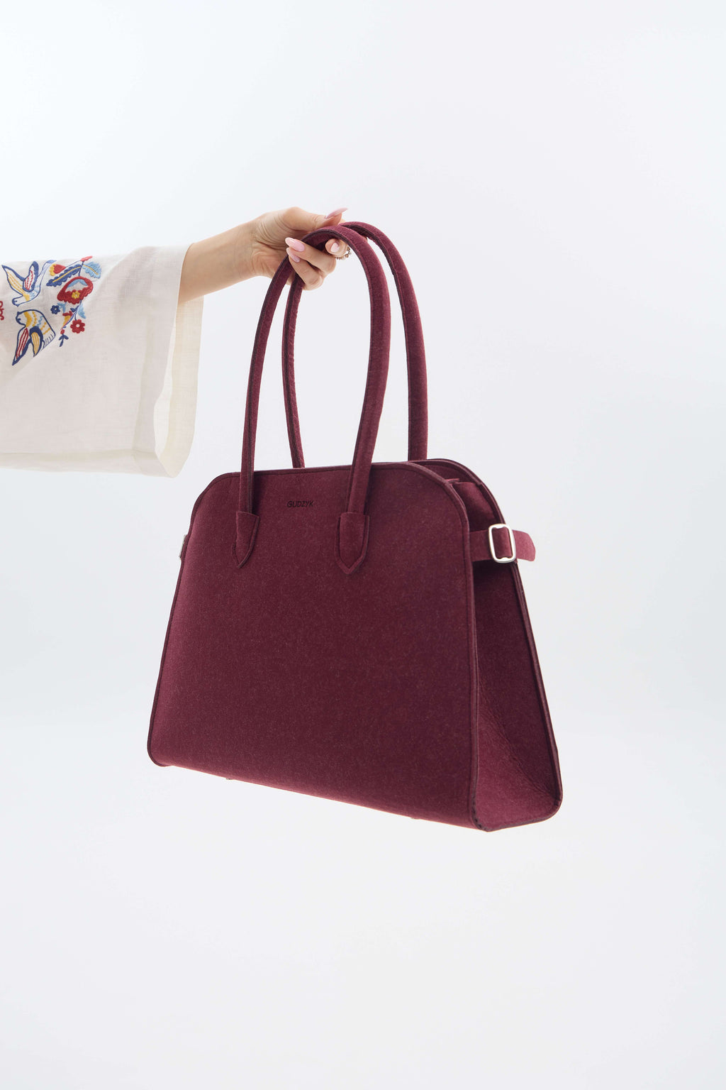Burgundy handbag held by a person with a white sleeve and colorful embroidery on a light gray background