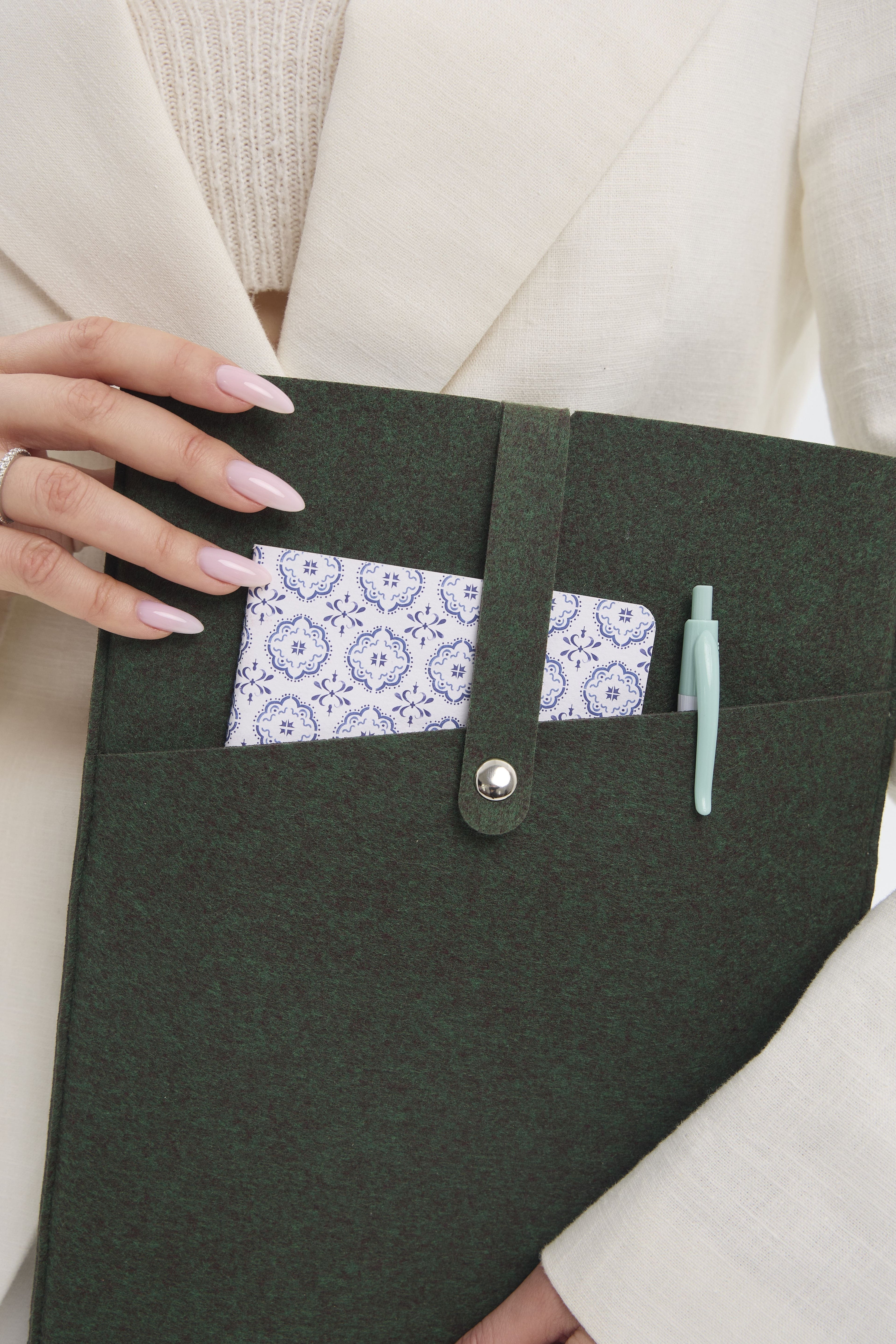 Dark green case with a pocket containing a white notebook and a pen, held by a person wearing a beige coat.