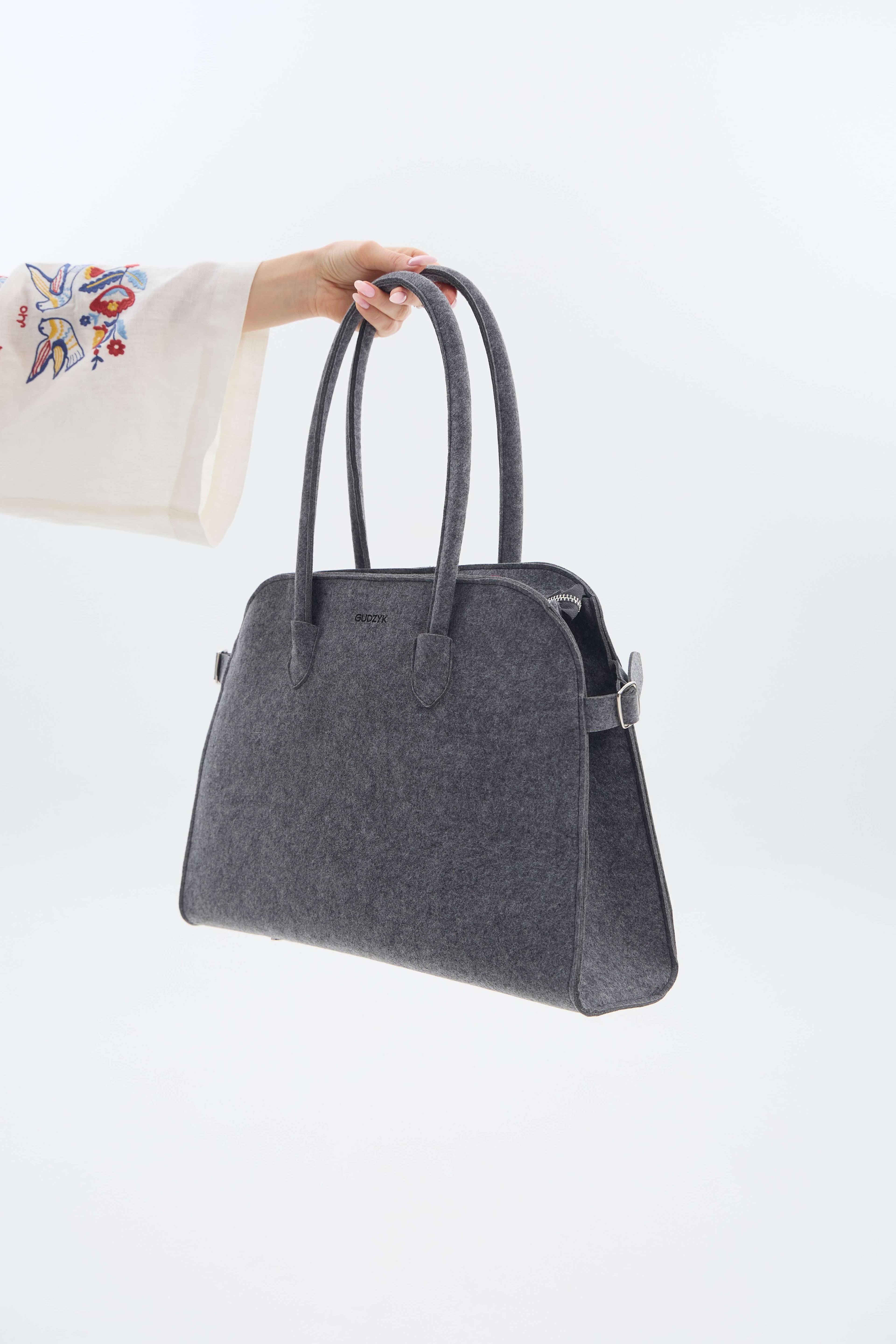 Gray handbag held by a person with a white sleeve and colorful embroidery on a light gray background