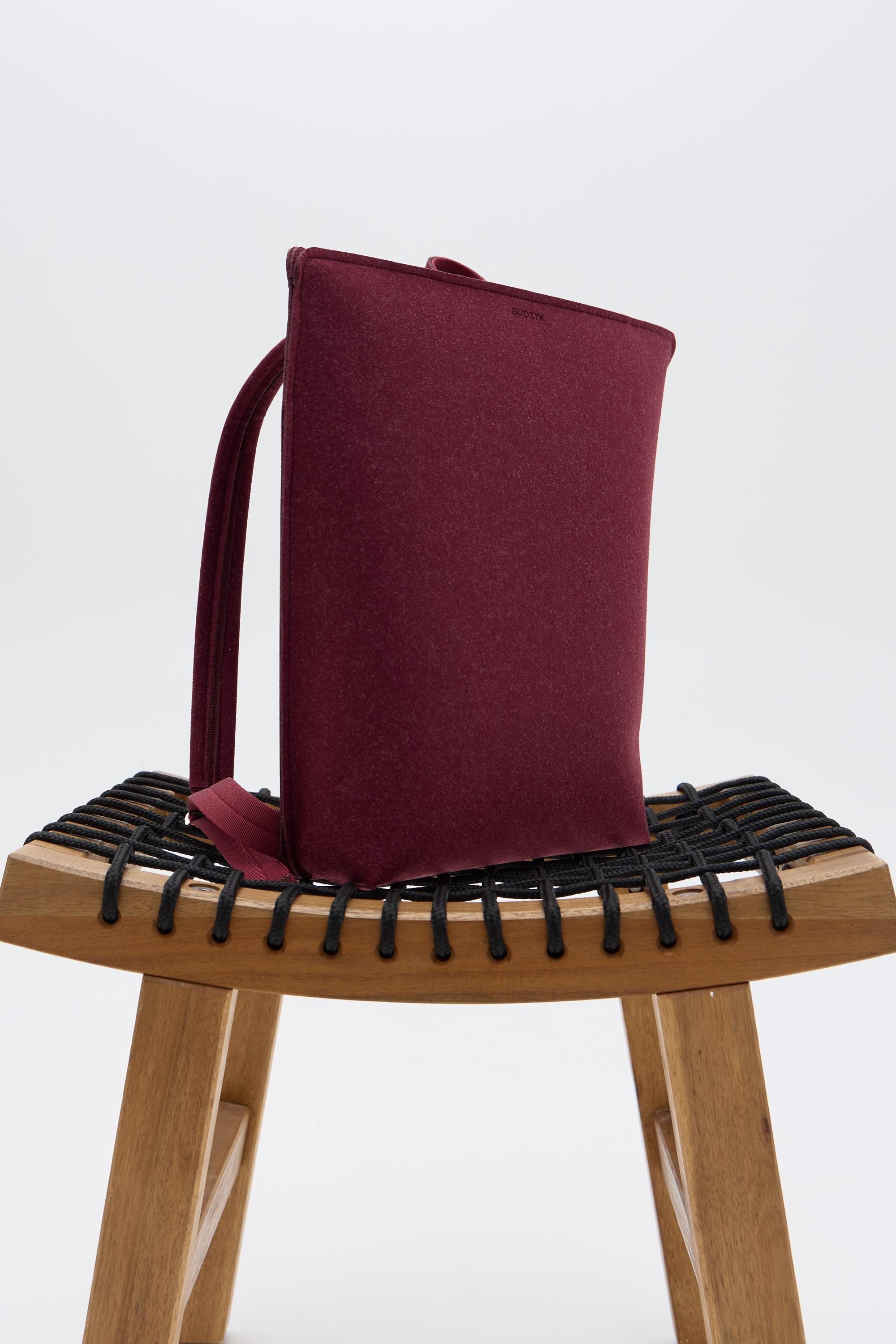 Maroon backpack on a wooden stool with black woven base against a white background