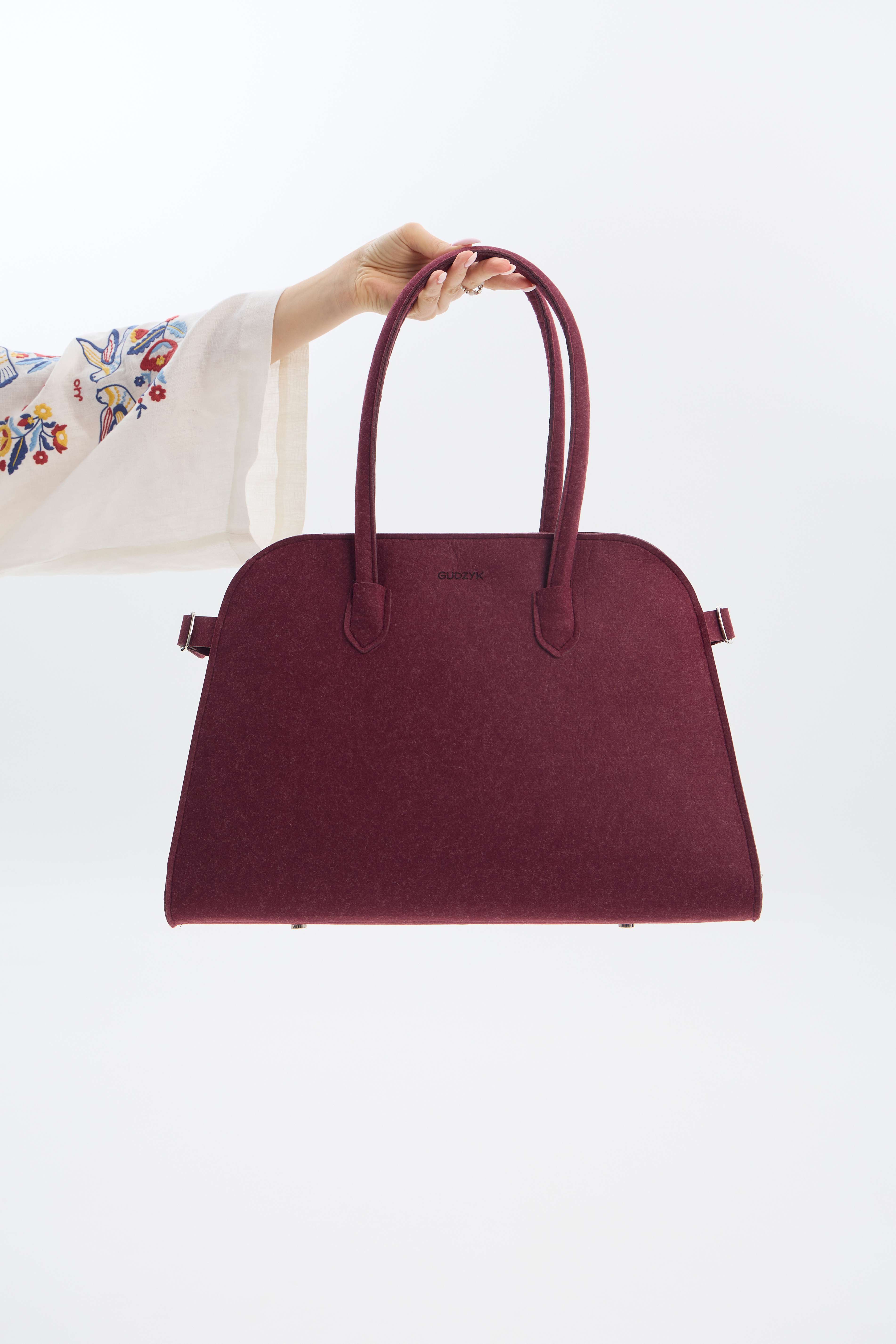 Maroon handbag held by a person with a white sleeve and colorful embroidery on a light gray background