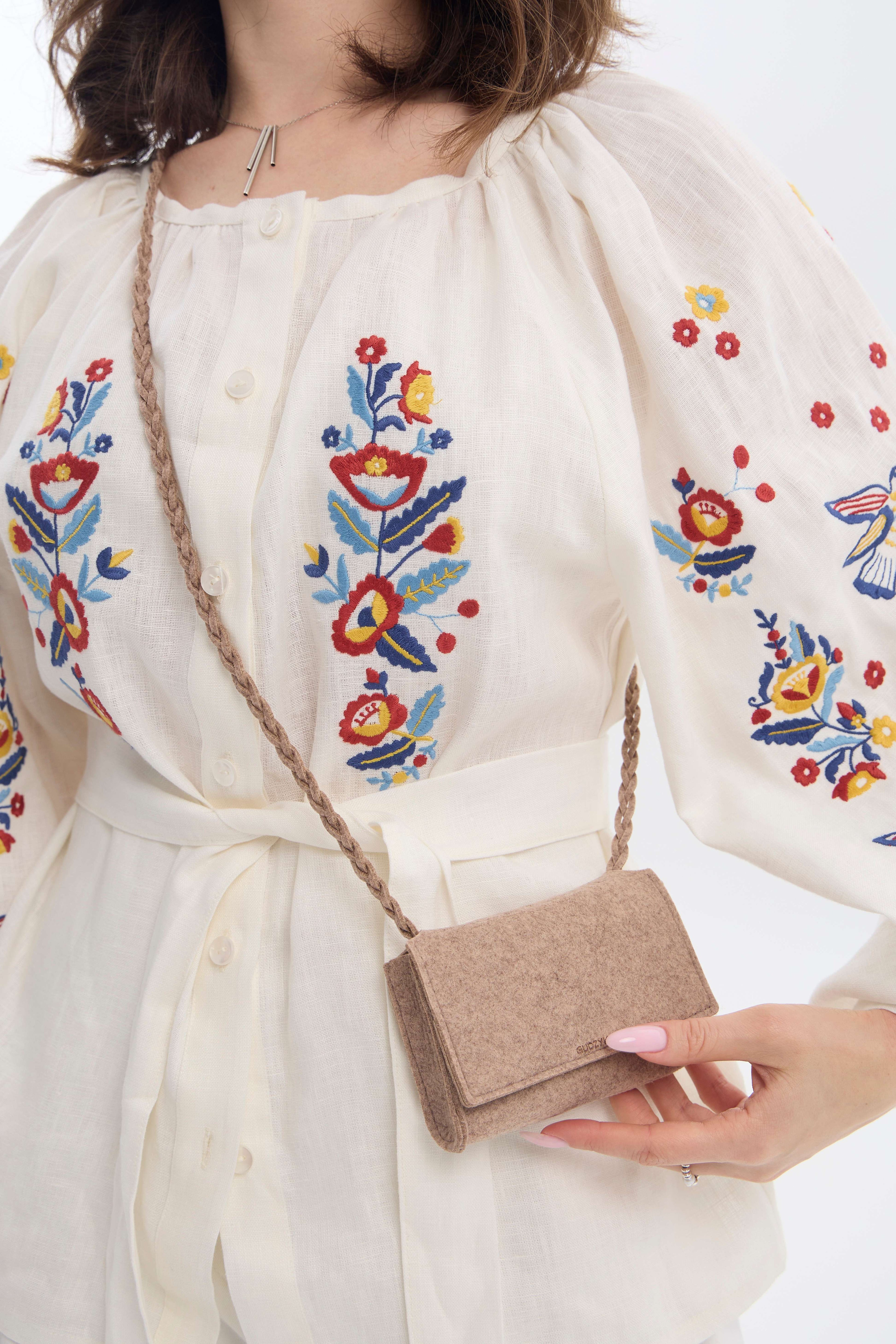 Person wearing a white dress with colorful floral embroidery holding a beige felt mini crossbody bag